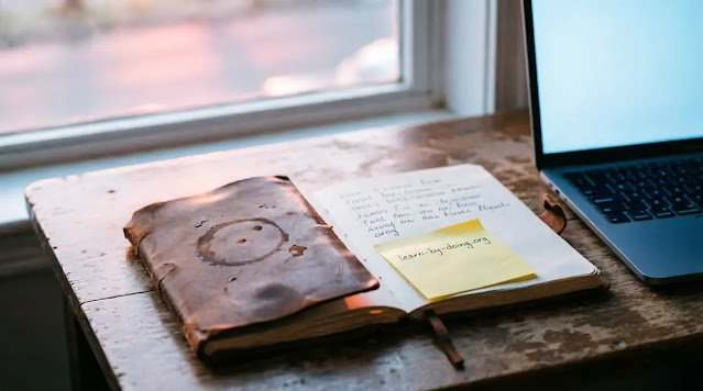 Coffee-stained notebook with first notes and URL sticky note beside laptop screen glowing at dawn window(AI-generated illustration)
