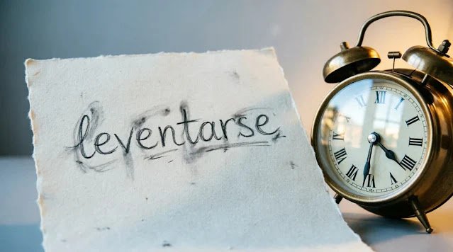 Word 'levantarse' handwritten with pencil smudges on cream paper beside vintage brass alarm clock with Roman numerals, soft gray-blue dawn light (AI-generated illustration)