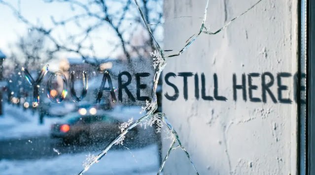 Shattered window glass with "YOU ARE STILL HERE" text written on surface (AI-generated illustration)