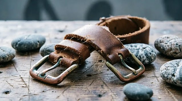 Leather strap loose on workbench, river stones scattered, brass buckle unfastened, soft light (AI-generated illustration)